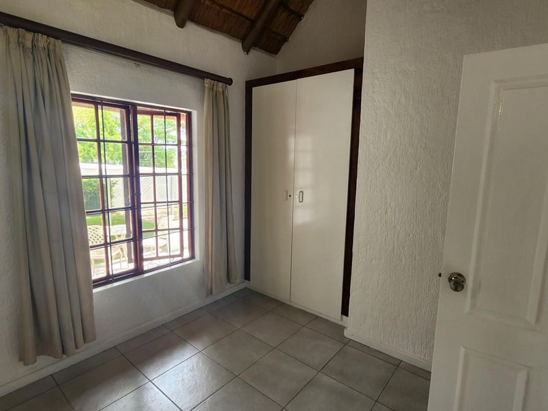 A bright, empty bedroom featuring tiled flooring, built-in cupboards, and a unique thatched ceiling with a timber-framed window overlooking the exterior.