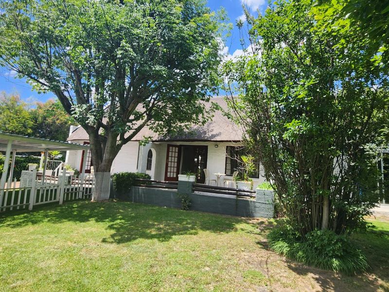 Rear view of a charming cottage featuring a traditional thatched roof, a covered patio area, and a well-maintained garden with mature trees.