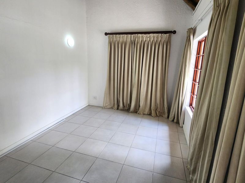 An empty bedroom featuring neutral tiled flooring, a wall-mounted light fixture, and a window with a wooden frame and curtain rod.