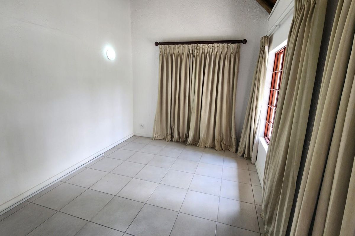 An empty bedroom featuring neutral tiled flooring, a wall-mounted light fixture, and a window with a wooden frame and curtain rod.