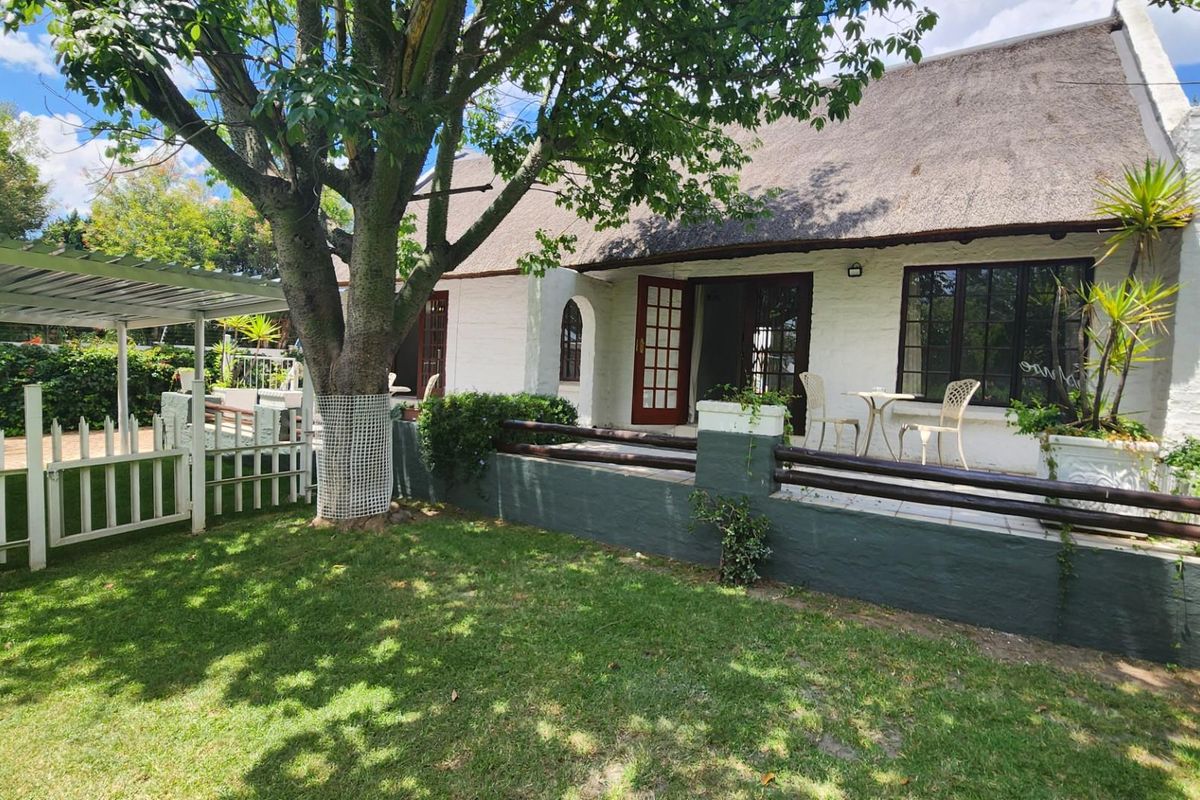 A charming cottage-style exterior featuring a traditional thatched roof, a raised patio area with timber railings, and a well-maintained garden space.