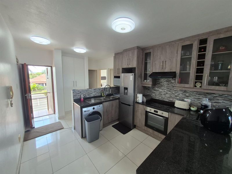 A functional kitchen featuring wood-look cabinetry, granite countertops, a built-in oven, extractor fan, and integrated laundry plumbing, with direct access to a balcony.