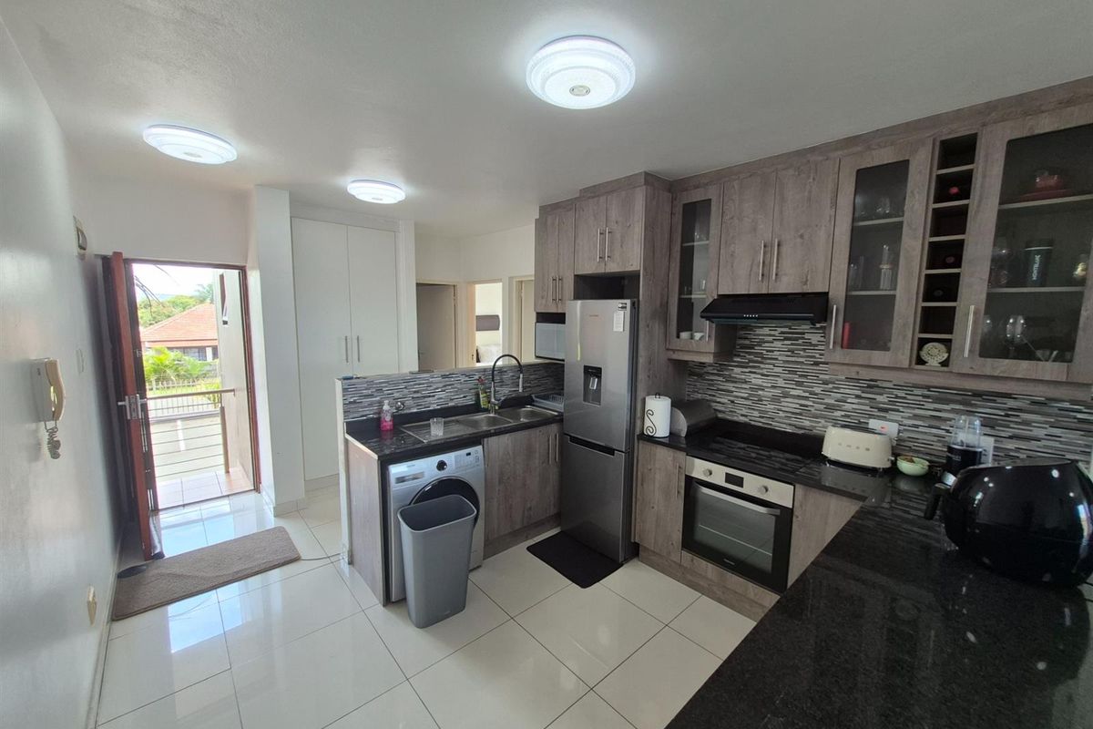 A functional kitchen featuring wood-look cabinetry, granite countertops, a built-in oven, extractor fan, and integrated laundry plumbing, with direct access to a balcony.