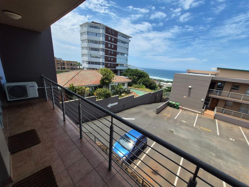 A balcony with tiled flooring and metal railings, offering an elevated view of the parking area, surrounding buildings, and the distant ocean.