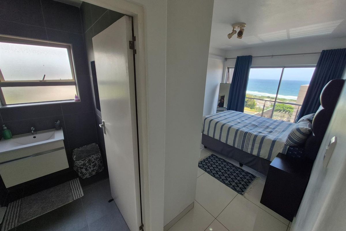 A view from the hallway looking into a modern bathroom with dark tiling and a bedroom featuring large windows with ocean views.