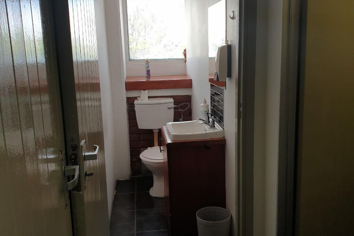 A compact industrial-style restroom featuring a built-in vanity, ceramic toilet, and dark floor tiling with natural light from a high window.
