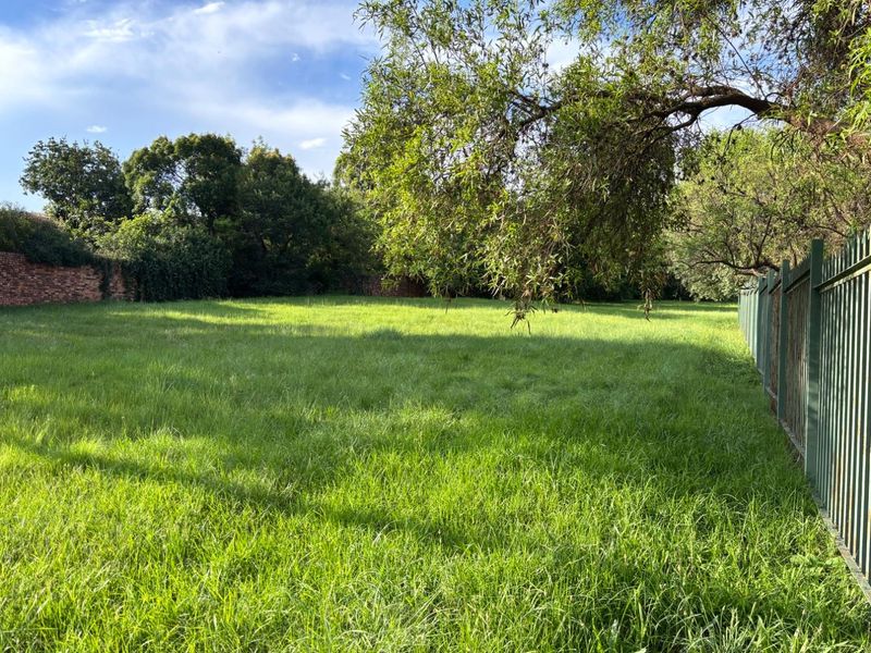 A spacious, level residential vacant plot featuring established trees, a brick boundary wall, and secure metal perimeter fencing.