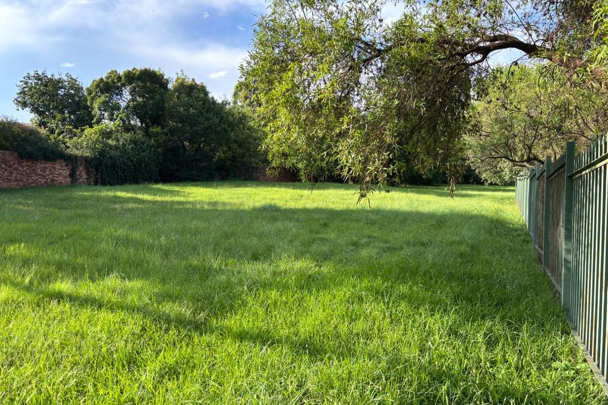 A spacious, level residential vacant plot featuring established trees, a brick boundary wall, and secure metal perimeter fencing.