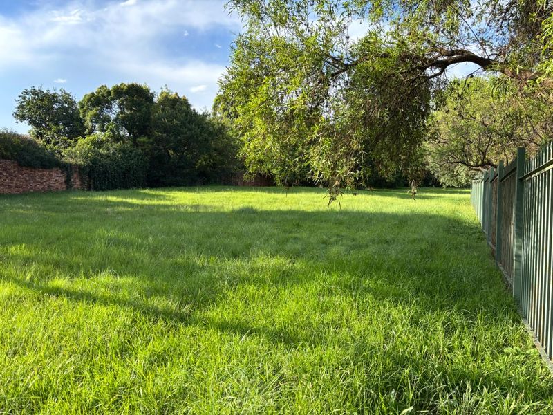 A spacious, level residential vacant land plot featuring established trees, a perimeter brick wall, and secure metal fencing.