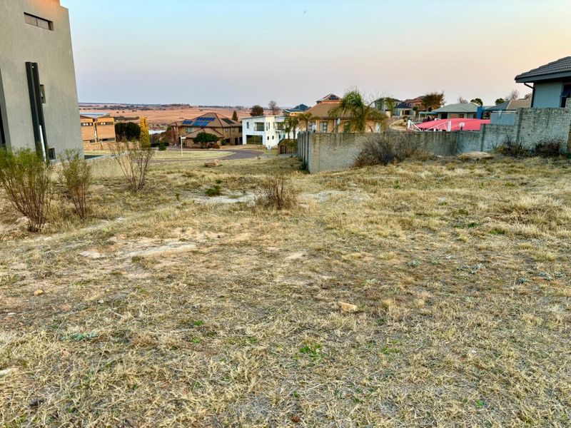 A view of a vacant residential plot featuring dry grass, surrounding boundary walls, and a view of the neighboring residential estate.