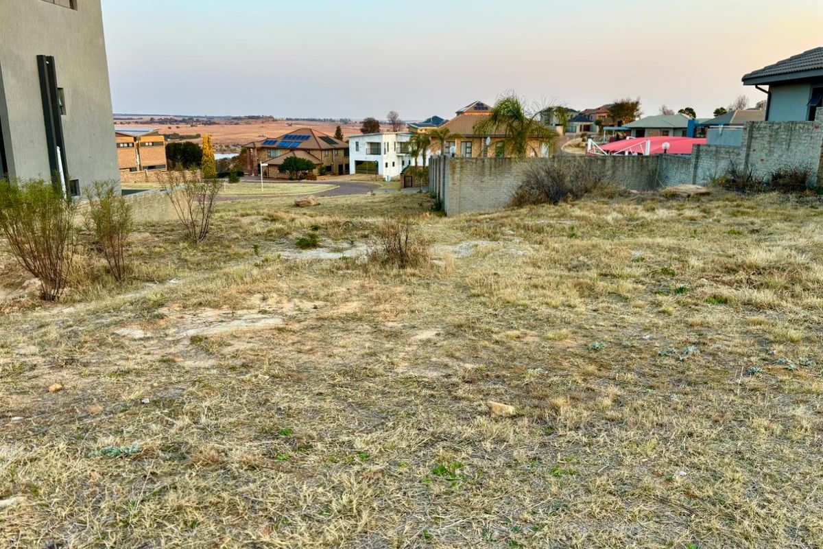 A view of a vacant residential plot featuring dry grass, surrounding boundary walls, and a view of the neighboring residential estate.
