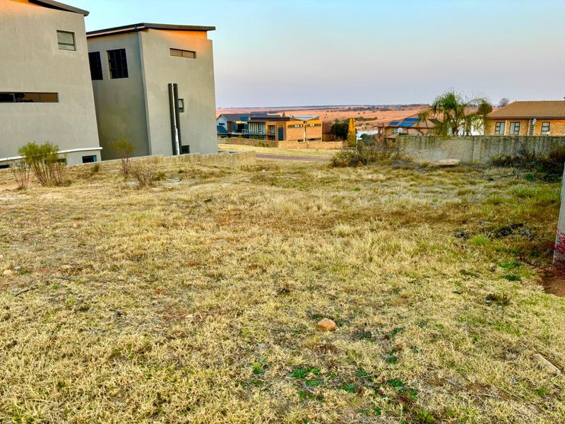 A vacant residential plot featuring dry grass, a boundary wall, and views of the surrounding neighborhood and landscape.