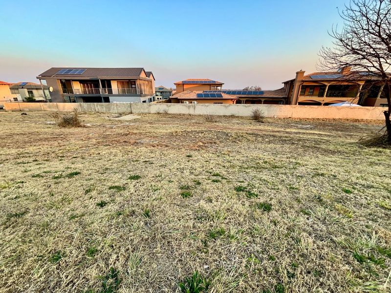 A view of a vacant residential plot featuring dry grass and a perimeter wall, with established neighboring homes visible in the background.