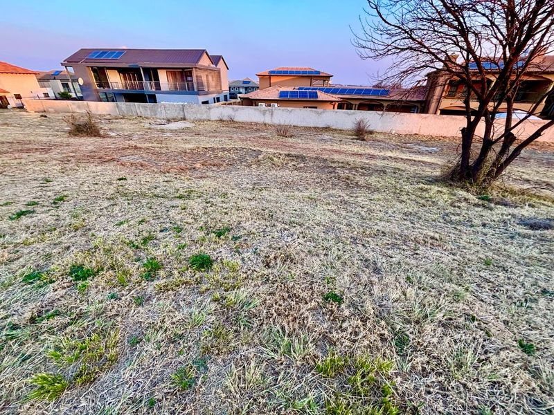 A view of a vacant residential plot featuring dry grass and a perimeter boundary wall, situated in a developed residential estate.