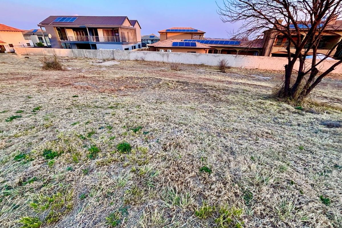 A view of a vacant residential plot featuring dry grass and a perimeter boundary wall, situated in a developed residential estate.