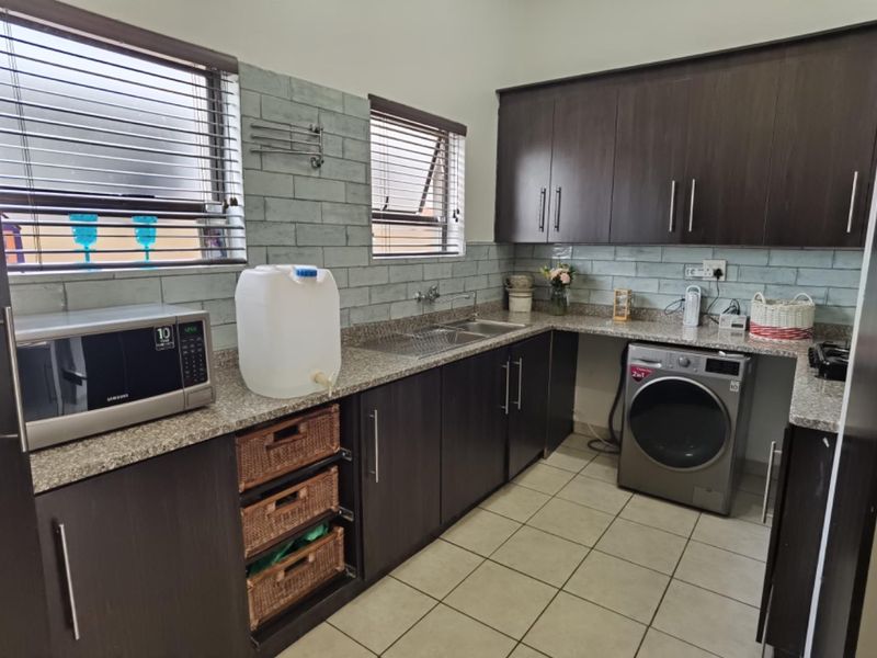 A functional scullery featuring dark wood cabinetry, granite countertops, a double basin sink, and tiled backsplash with integrated laundry appliance space.