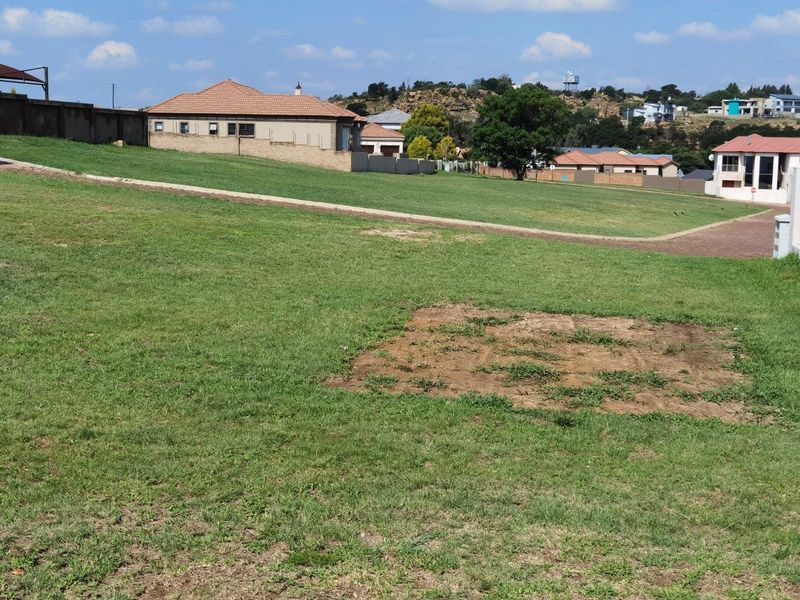 A vacant residential plot featuring a grass lawn, paved driveway access, and surrounding boundary walls in a developed neighborhood.