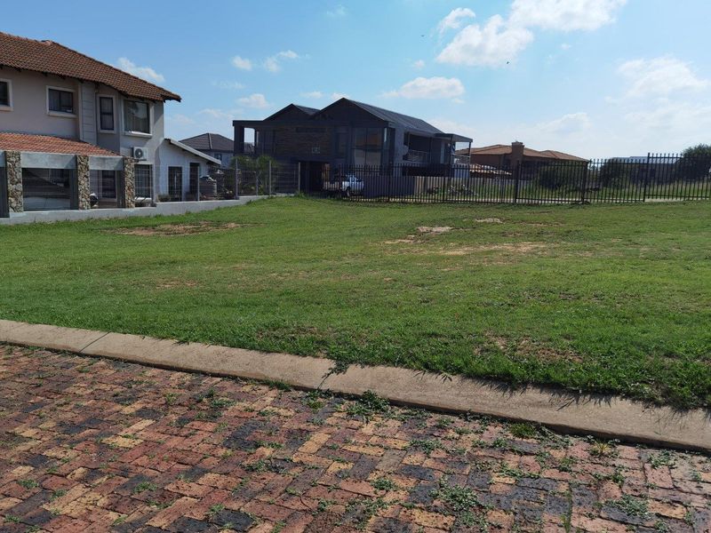 A vacant residential plot featuring a grass lawn, paved driveway, and metal perimeter fencing in a suburban neighborhood setting.
