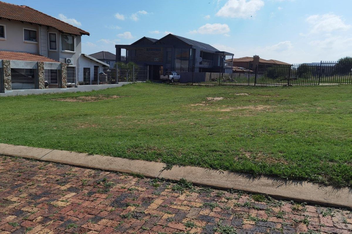 A vacant residential plot featuring a grass lawn, paved driveway, and metal perimeter fencing in a suburban neighborhood setting.