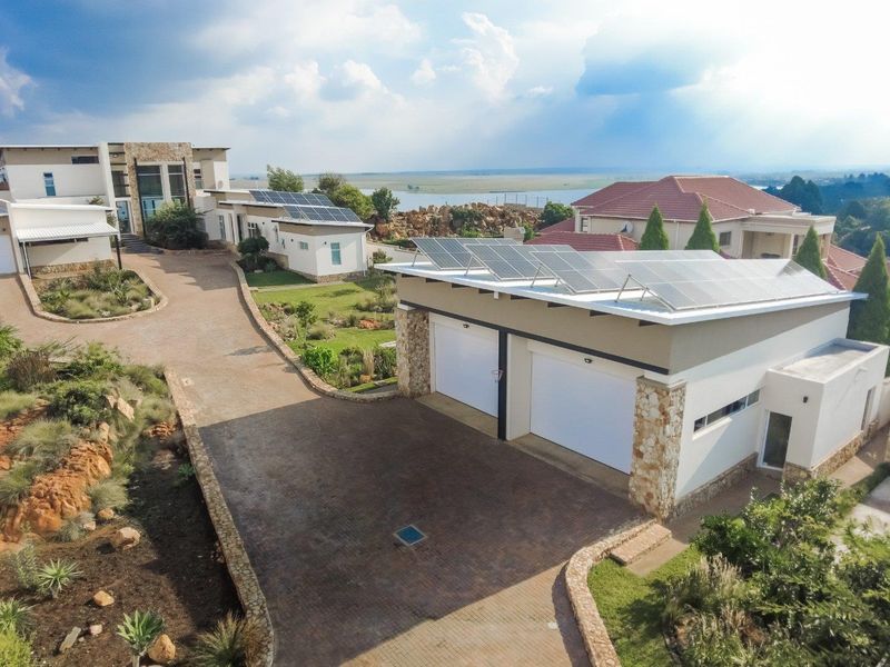 Aerial view of a modern property featuring a large paved driveway, double garage with solar panels, and landscaped grounds with scenic water views.