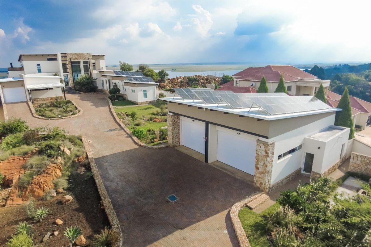 Aerial view of a modern property featuring a large paved driveway, double garage with solar panels, and landscaped grounds with scenic water views.
