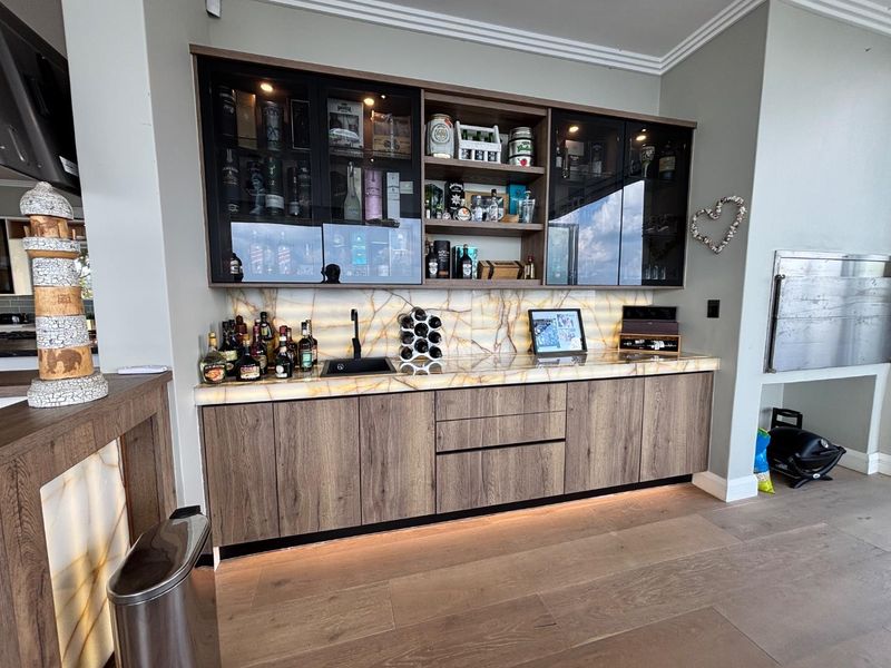 A modern, high-end built-in bar area featuring custom wood cabinetry, a striking backlit stone countertop, integrated display shelving, and a sleek undermount sink.