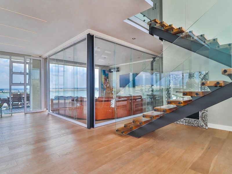 Modern open-plan living area featuring a striking floating staircase with glass balustrades and a unique glass-enclosed room overlooking the exterior deck and water view.