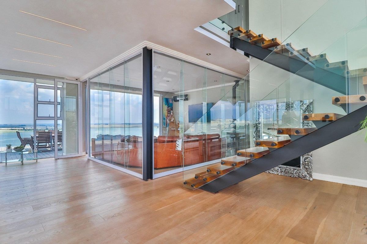 Modern open-plan living area featuring a striking floating staircase with glass balustrades and a unique glass-enclosed room overlooking the exterior deck and water view.