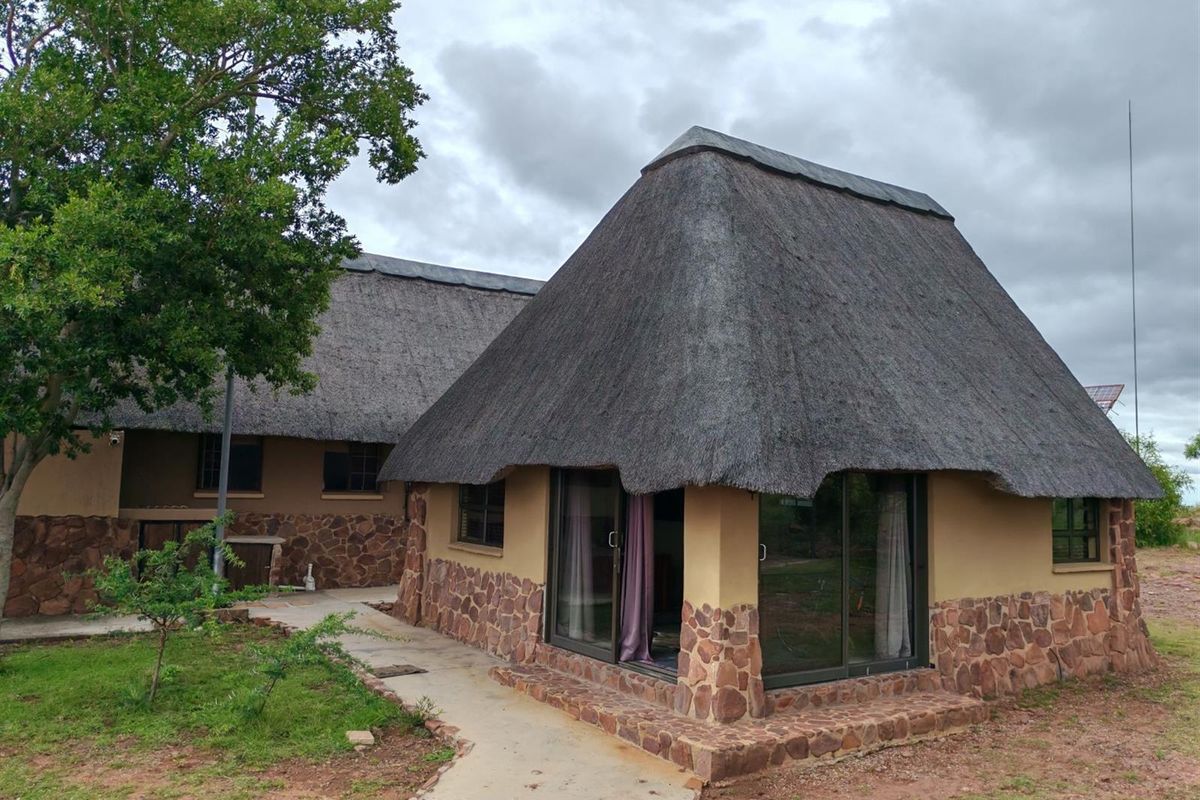 A rustic exterior view of a thatched-roof dwelling featuring stone cladding and sliding glass doors, set within a natural garden landscape.