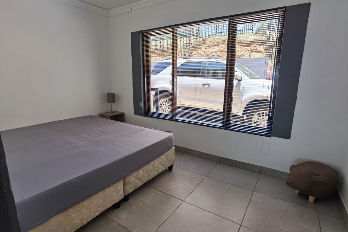 A bedroom featuring large windows with venetian blinds and neutral tiled flooring, offering a functional space with natural light.
