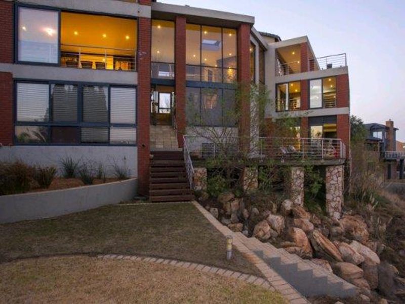 A multi-level rear elevation featuring extensive glazing, a raised timber deck, and stone-clad structural pillars overlooking a landscaped garden and rocky terrain.