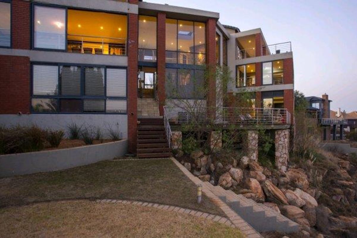 A multi-level rear elevation featuring extensive glazing, a raised timber deck, and stone-clad structural pillars overlooking a landscaped garden and rocky terrain.