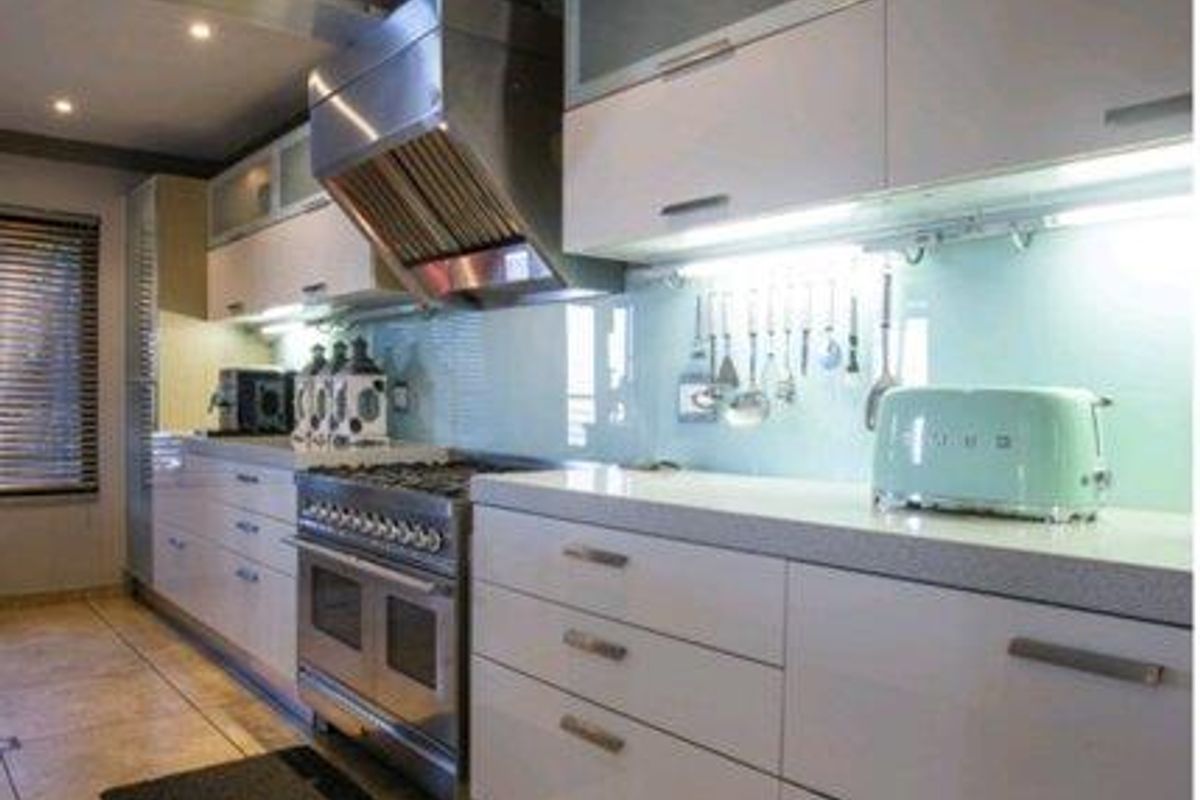 Modern kitchen featuring sleek white cabinetry, stone countertops, a stainless steel gas range cooker, and a glass splashback with integrated under-cabinet lighting.