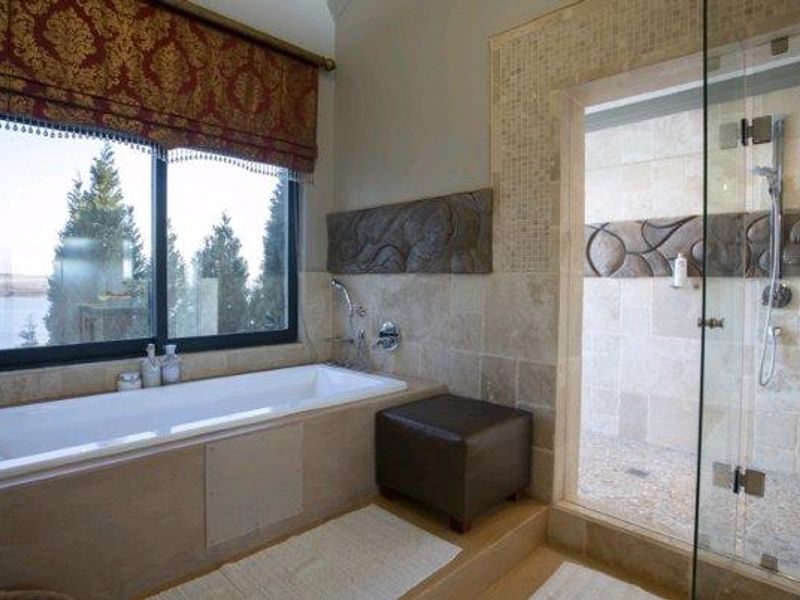 A well-appointed bathroom featuring a built-in bathtub, a spacious walk-in shower with glass enclosure, and elegant travertine wall tiling with decorative stone accents.