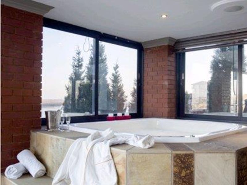 A luxurious bathroom featuring a raised built-in bathtub with stone tile surround, exposed brick accents, and large windows offering scenic outdoor views.