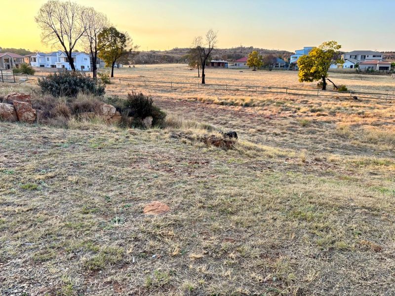 A wide-angle view of a vacant residential plot featuring natural grass, scattered trees, and perimeter fencing with surrounding neighborhood houses in the distance.