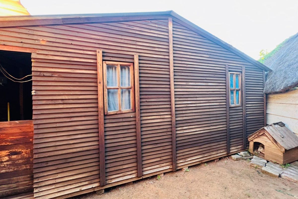 Exterior view of a timber-clad outbuilding featuring two small windows and a side entrance, showing signs of weathering and age.
