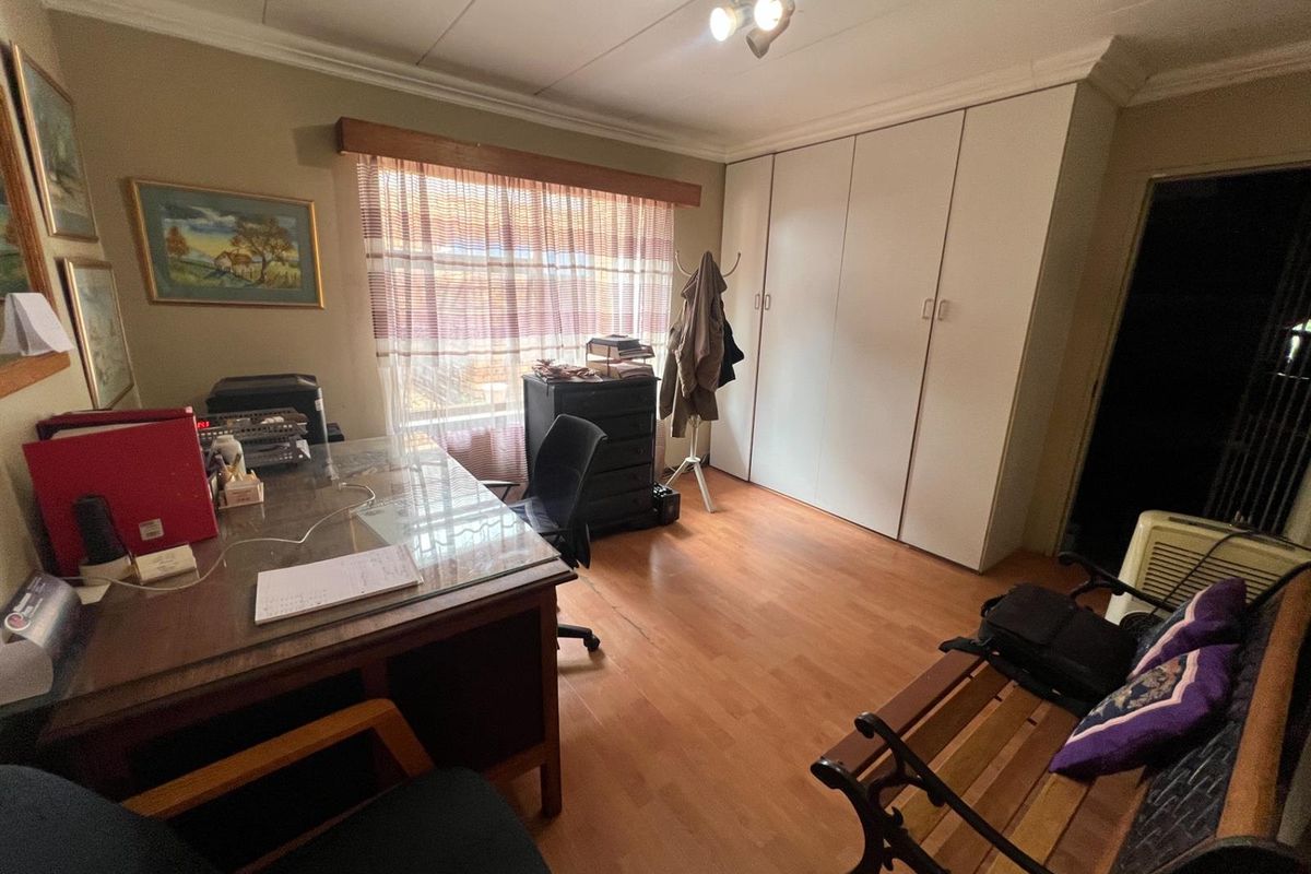 A functional bedroom featuring built-in cupboards, laminate flooring, and a window with curtaining, currently utilized as a home office space.