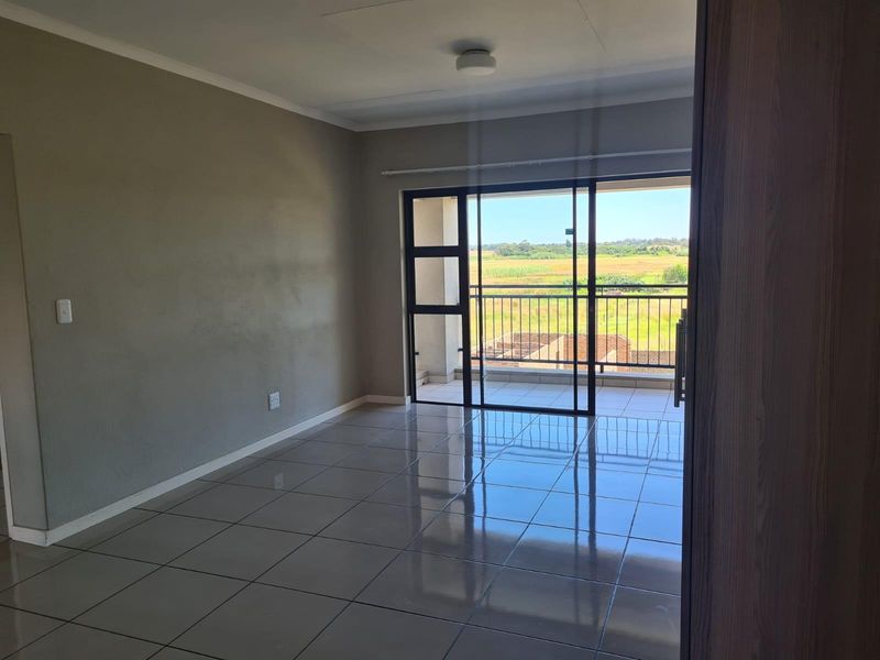 An empty, well-lit living area featuring neutral tiled floors and a large sliding glass door providing access to a balcony with scenic views.
