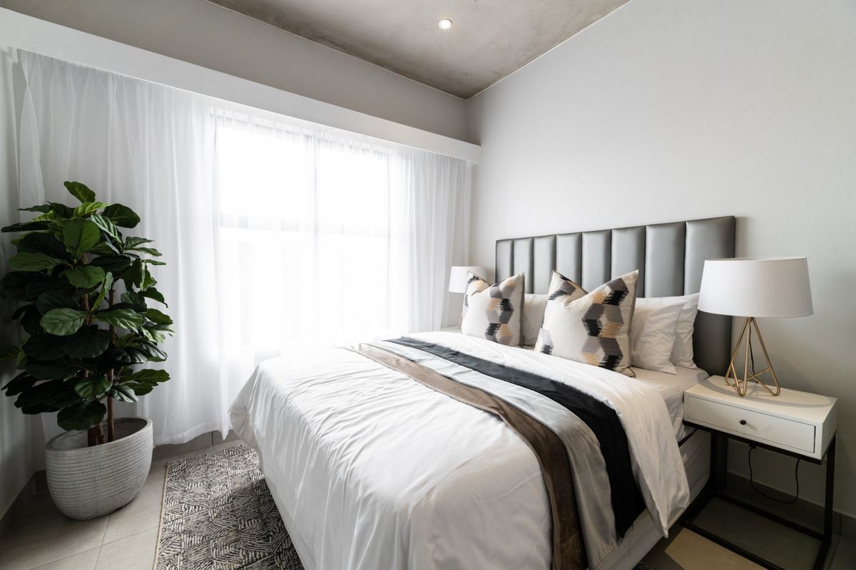 A modern bedroom featuring a neutral color palette, exposed concrete ceiling, and large window with sheer curtains.