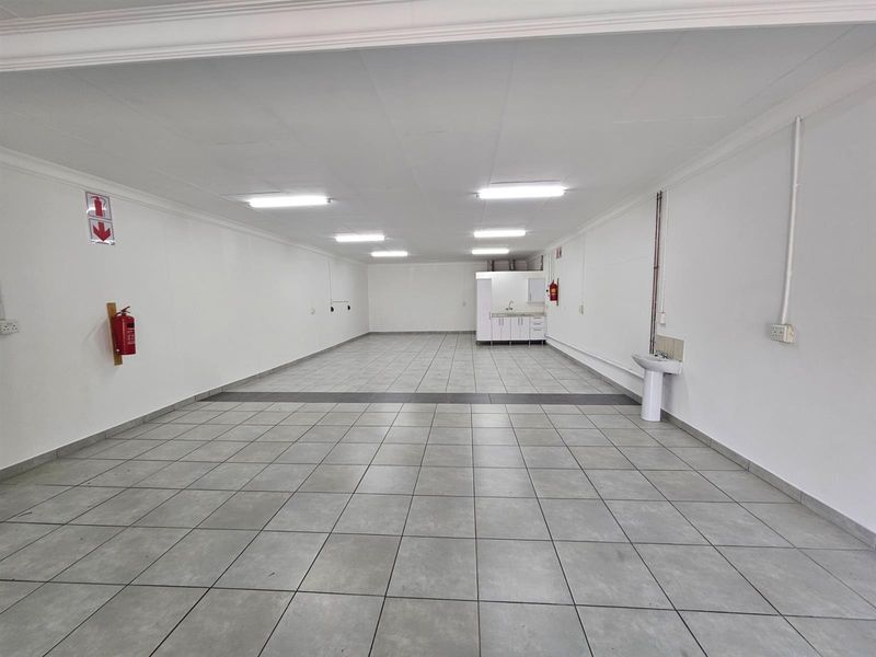 Spacious open-plan office area featuring tiled flooring, recessed fluorescent lighting, a small kitchenette corner, and a wall-mounted hand basin.