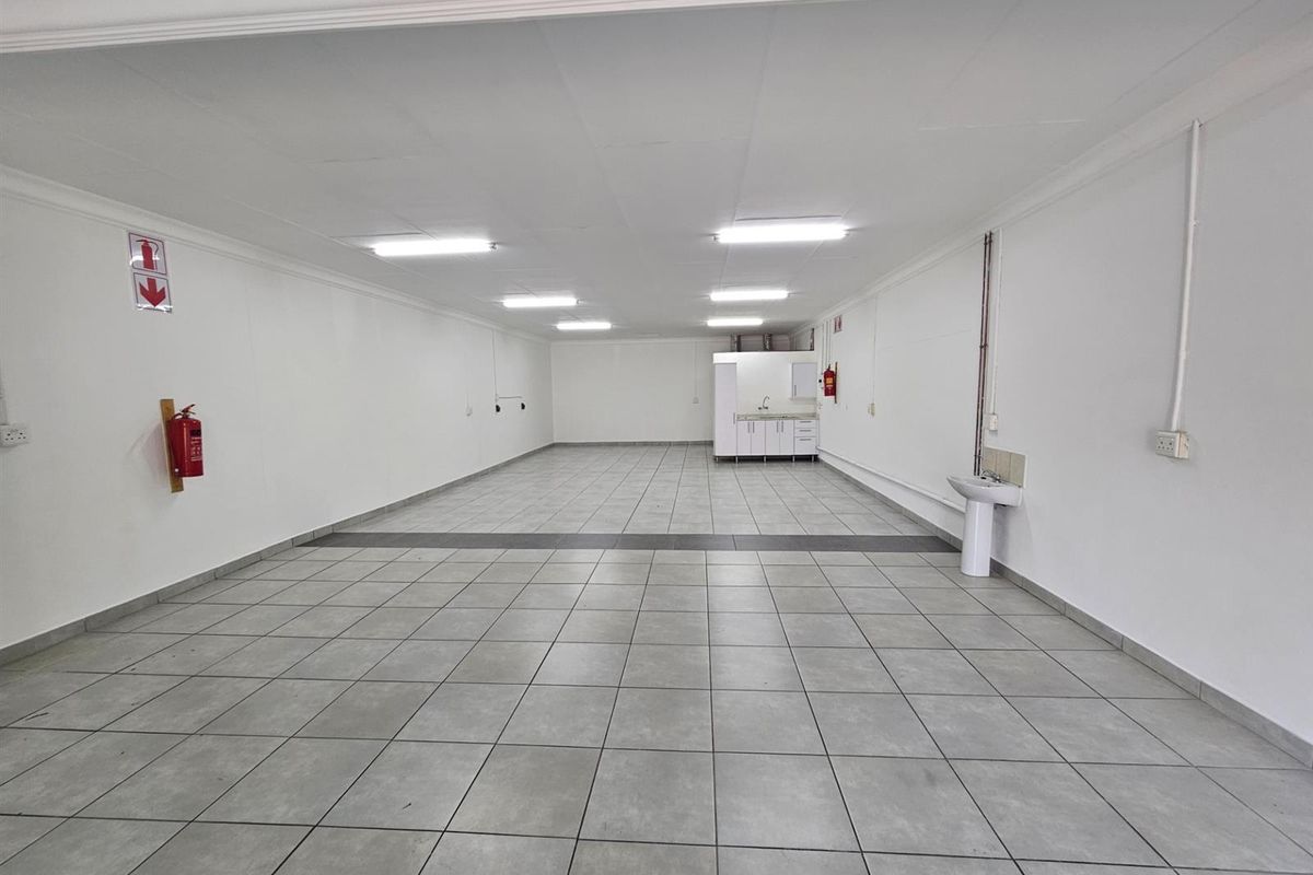Spacious open-plan office area featuring tiled flooring, recessed fluorescent lighting, a small kitchenette corner, and a wall-mounted hand basin.
