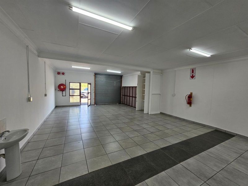Open plan office space featuring tiled flooring, fluorescent lighting, a built-in washbasin, roller shutter door, and wall-mounted fire safety equipment.