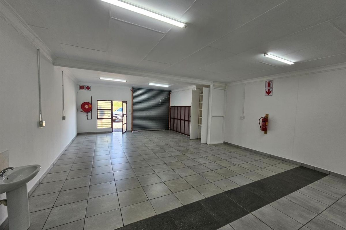 Open plan office space featuring tiled flooring, fluorescent lighting, a built-in washbasin, roller shutter door, and wall-mounted fire safety equipment.