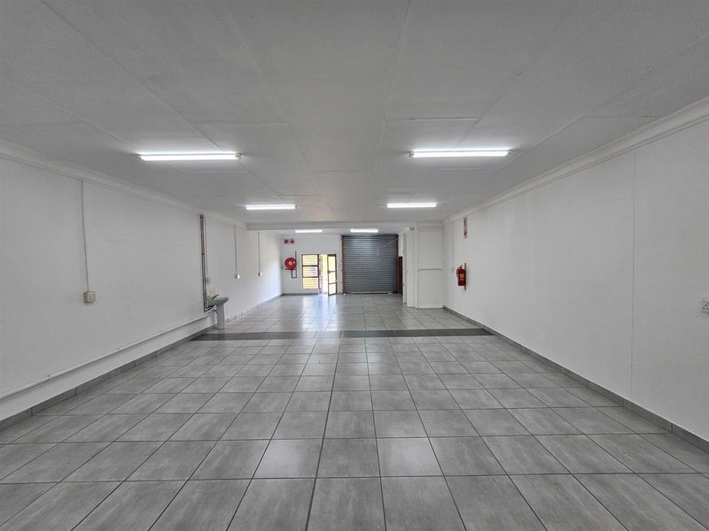 Spacious open-plan office area featuring tiled flooring, recessed fluorescent lighting, a small wall-mounted basin, and a secure roller shutter door entrance.