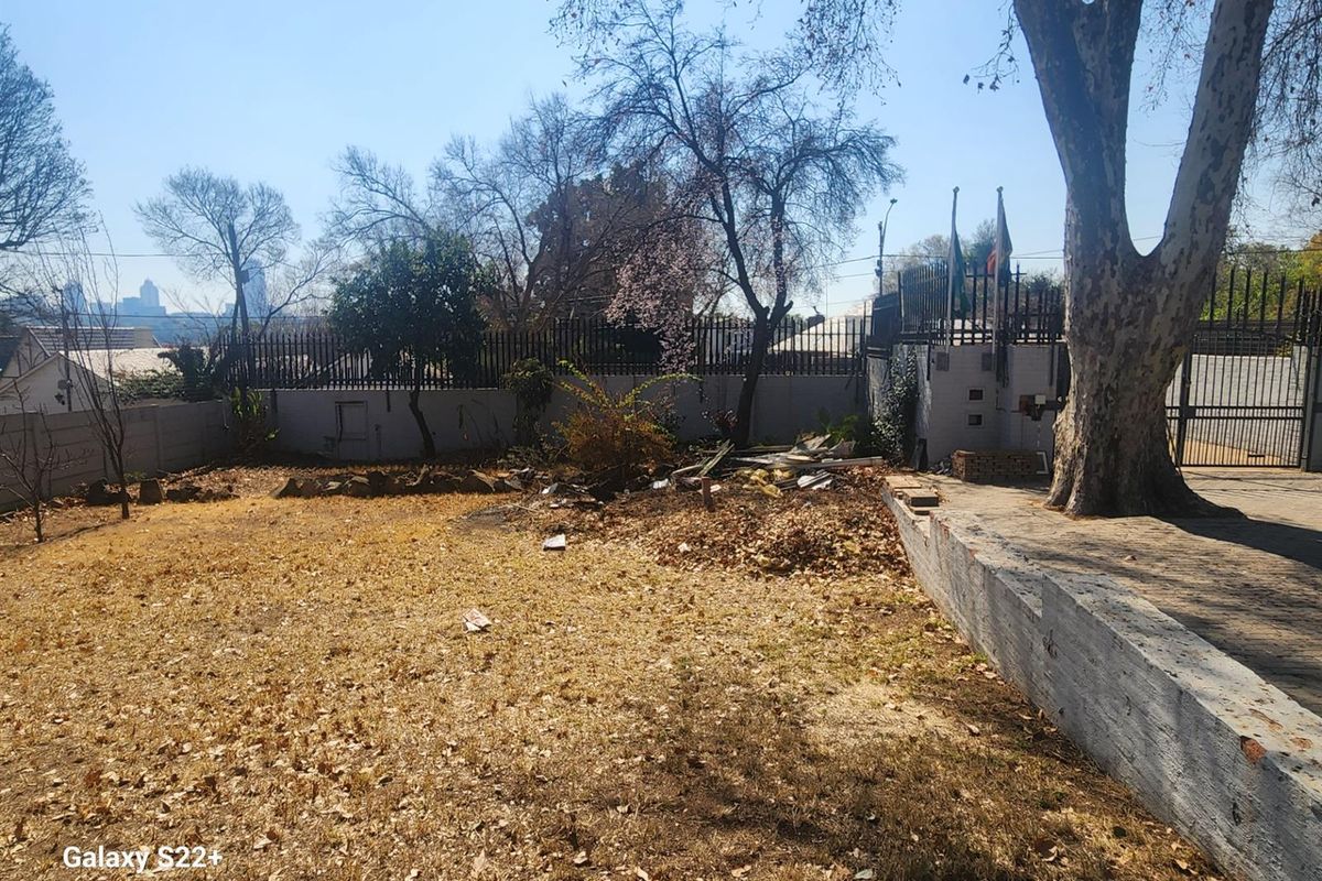 A spacious rear garden area featuring a retaining wall, mature trees, and perimeter security fencing, requiring landscaping and maintenance.