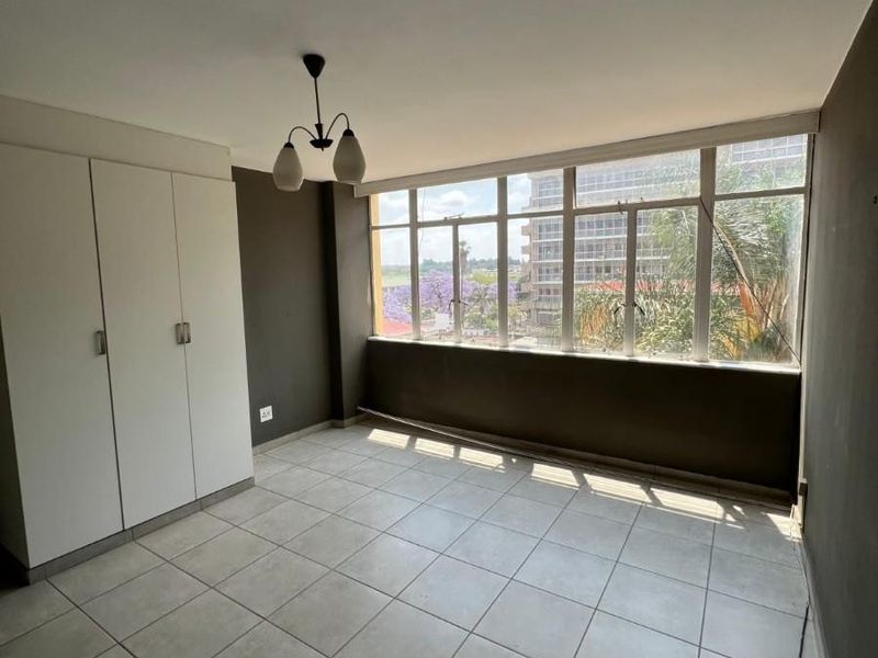 A bright, empty bedroom featuring tiled flooring, a built-in wardrobe, and large windows offering an urban view.