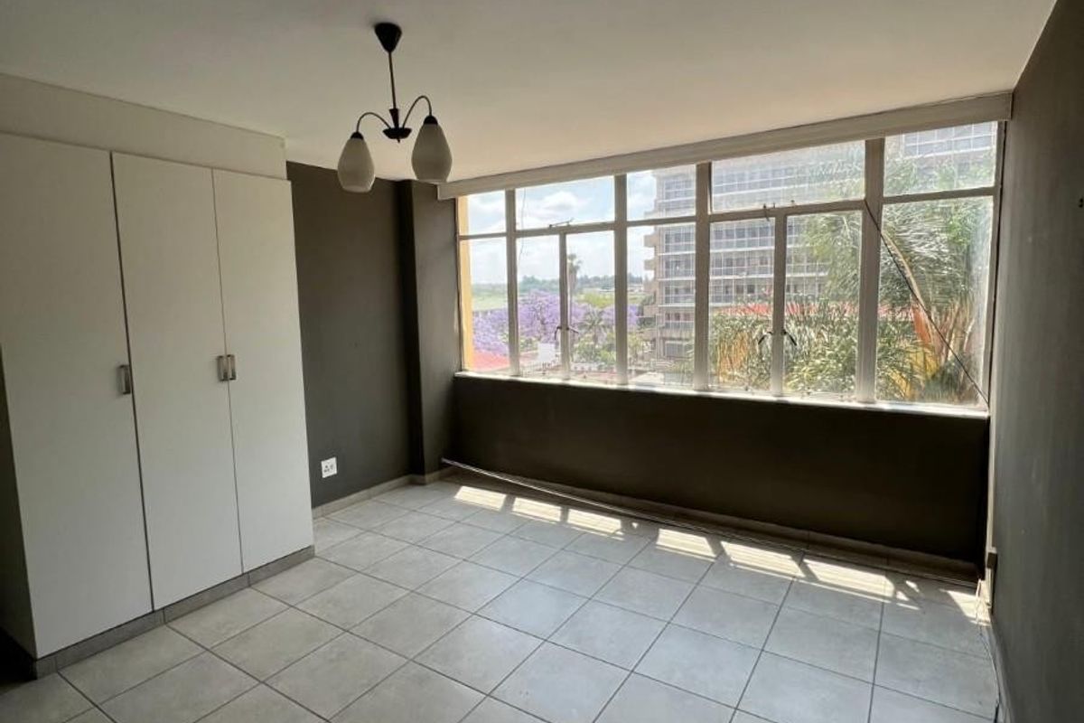 A bright, empty bedroom featuring tiled flooring, a built-in wardrobe, and large windows offering an urban view.