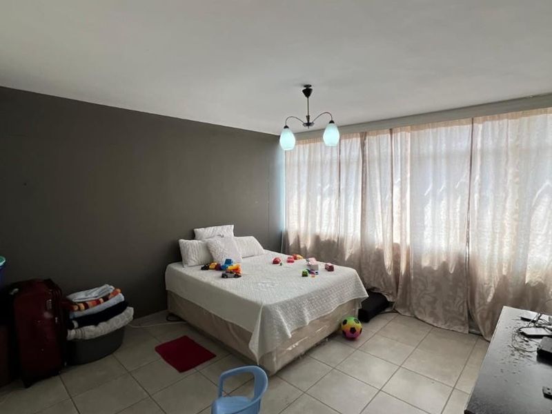 A spacious bedroom featuring neutral tiled flooring, a large window with curtains, and a central ceiling light fixture.