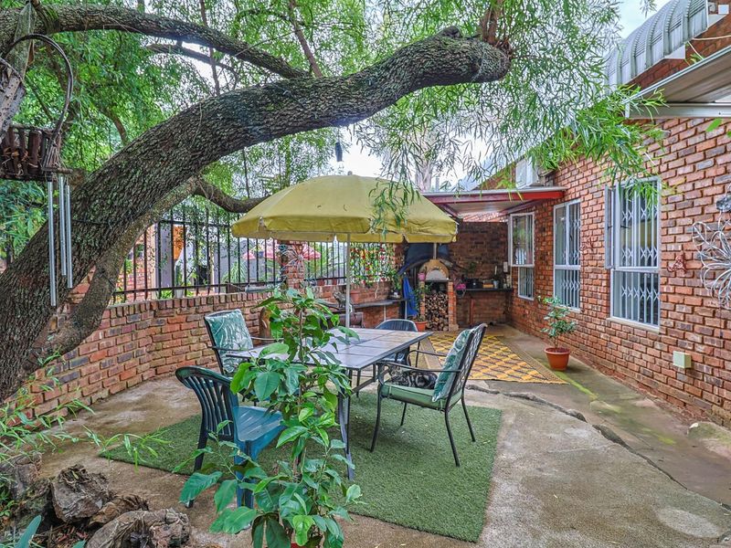 A covered outdoor patio area featuring exposed brick walls, concrete paving, and a built-in pizza oven, shaded by a large mature tree.